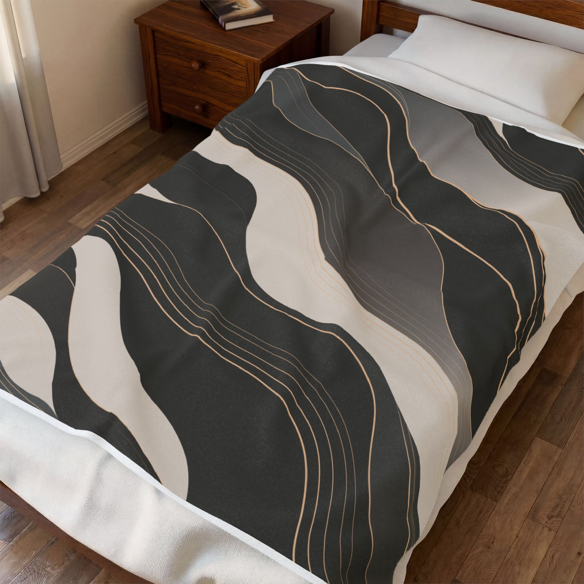 A bed in a bedroom setting with a modern abstract wavy patterned throw blanket. The blanket features large organic shapes in charcoal gray, off-white, and medium gray, accented by thin gold lines.