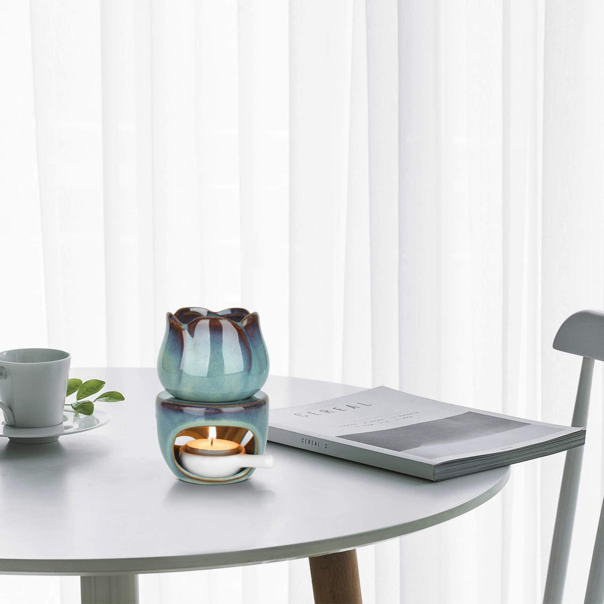VibraMist Eternal Bloom fragrance warmer with a lit tea light on a white table beside a cup and open book, against sheer curtains.
